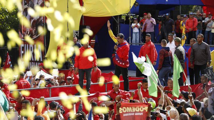 President Maduro stands on a stage waving, surrounded by supporters waving flags and placards